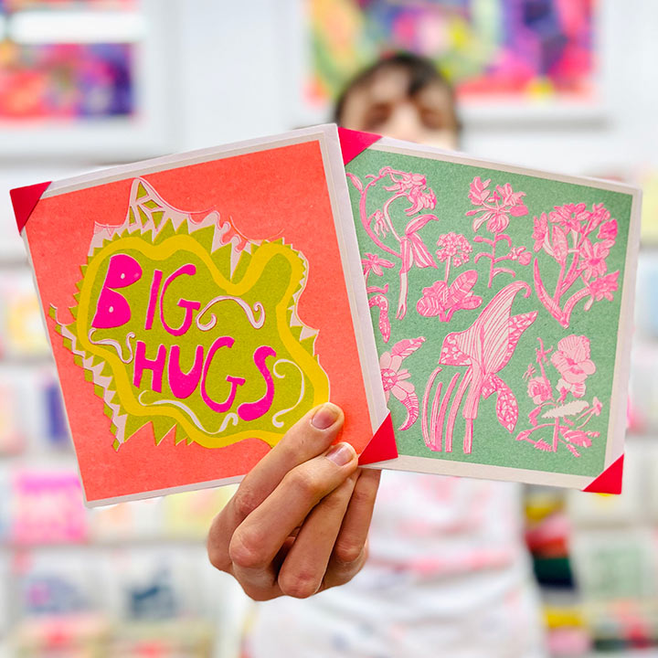 An artist holding 2 cards focusing on An orange, green and pink hand drawn card with the words big hugs