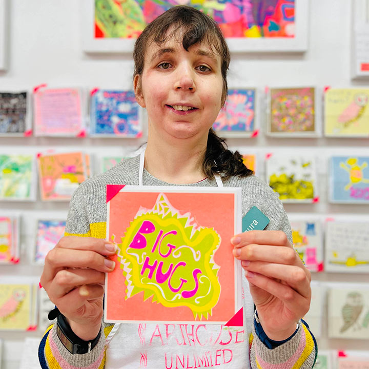 A female artist holding An orange, green and pink hand drawn card with the words big hugs