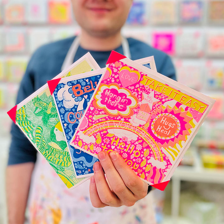 An artist holding 3 cards focusing on A bright pink, orange and yellow card with positive messages including the words take it easy