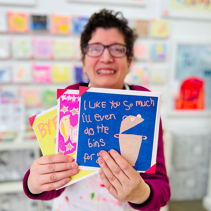 A female artist holding 3 cards focusing on A blue and orange drawing of a bin and the words I like you so much I'll even do the bin for you