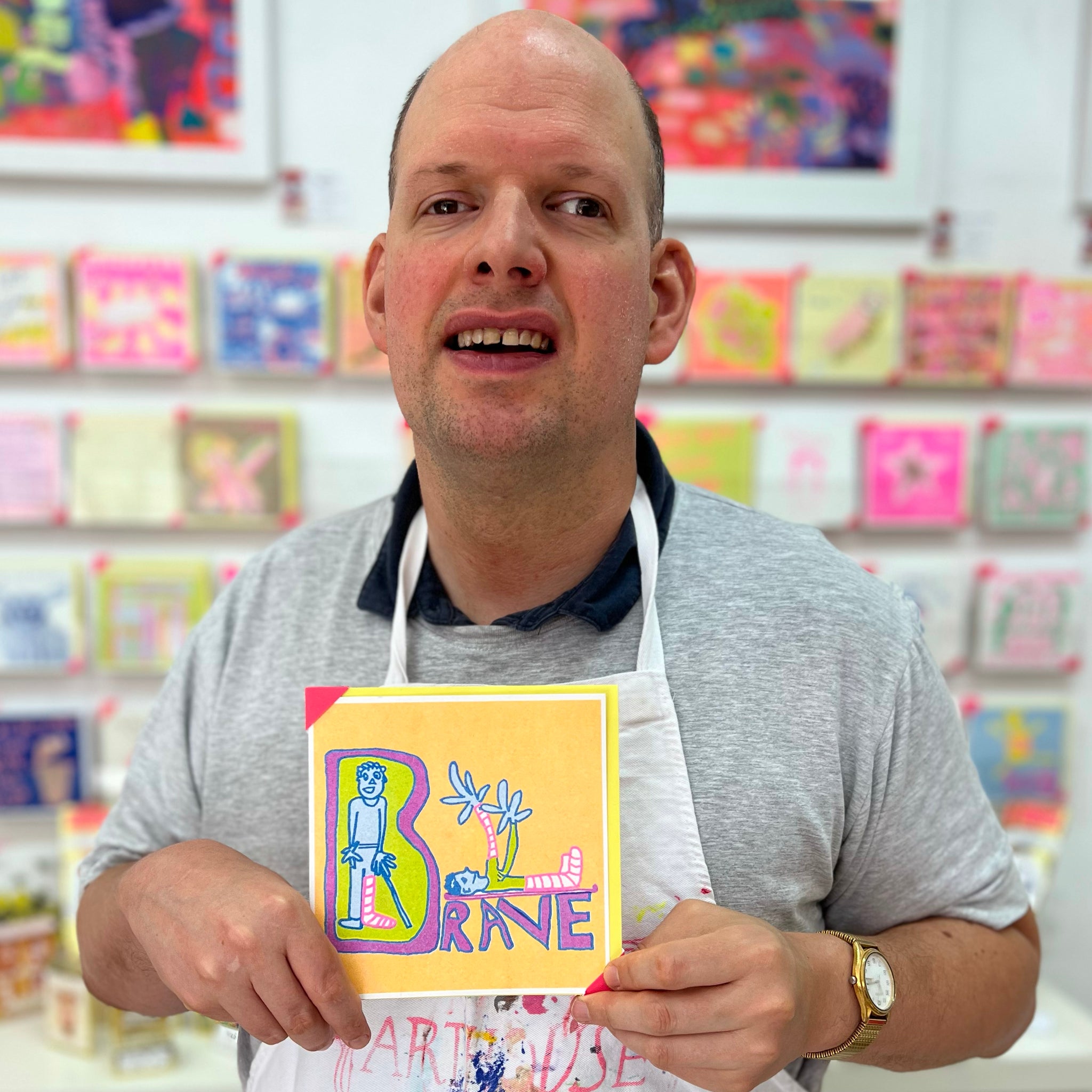A male artist holding An orange, blue and purple card with two hand drawn characters with the word brave