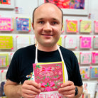 A male artist holding A pink, green and copper coloured hand drawn card with bows and christmas bells
