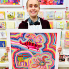 Male artist holding Framed painting of bright coloured strange creatures 