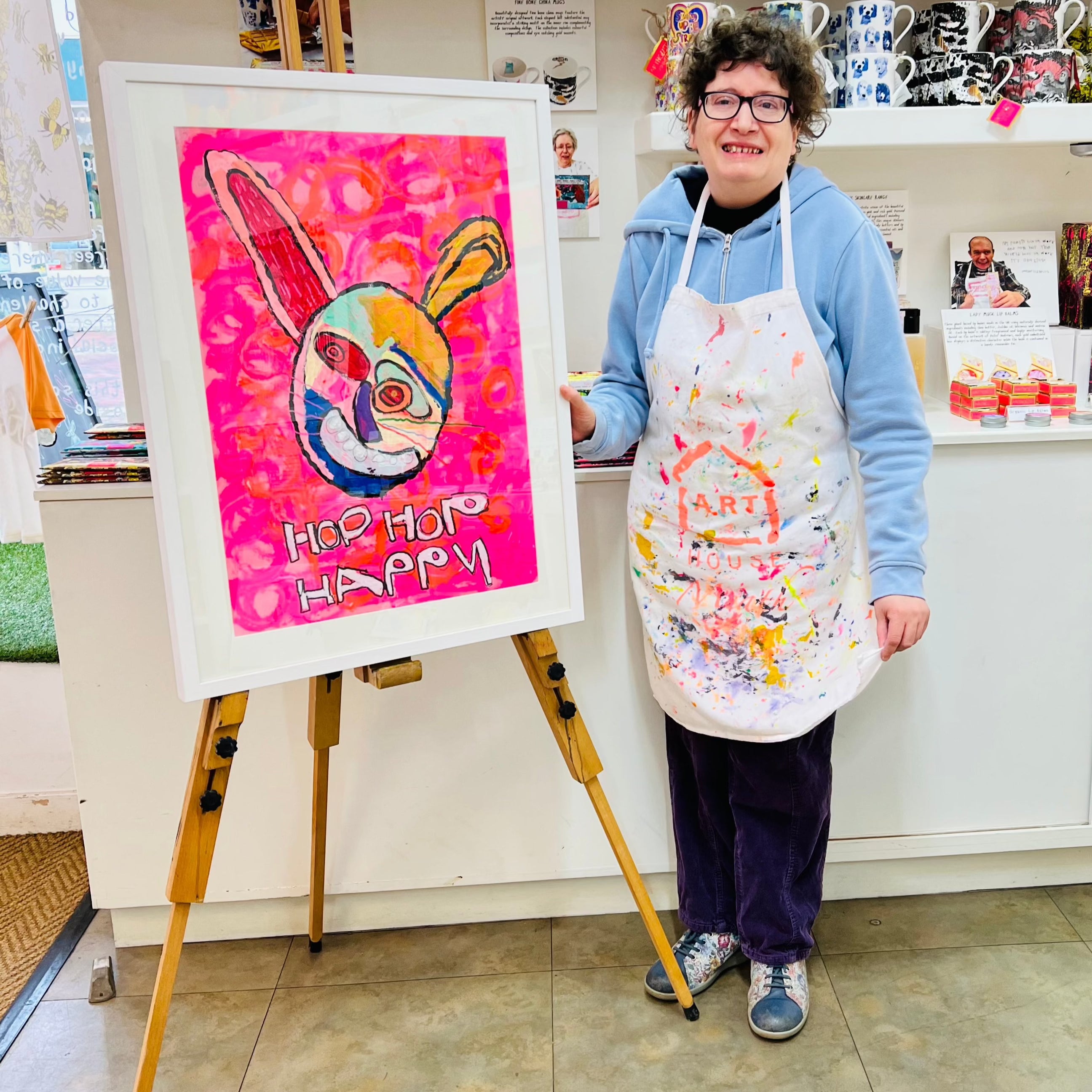 Female artist holding Framed painting of a bright coloured rabbit in pinks, red, green and orange with the words hop hop happy